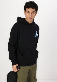 Black hoodie with drawstrings, front pocket, and blue mountain logo on the chest. Worn with olive green cargo pants and holding a black bag.