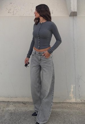 Kobieta stoi oparta o popękany betonowy mur, ubrana w dopasowaną, szarą, zapinaną na guziki krótką bluzkę, luźne, jasnoszare jeansy, trzyma okulary przeciwsłoneczne w jednej ręce.