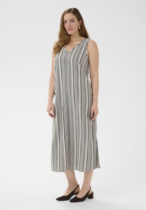 Kobieta stojąca w sukience bez rękawów sięgającej kostek, w pionowe szare i białe paski, oraz w czarne sandały na niskim obcasie z odkrytymi piętami.