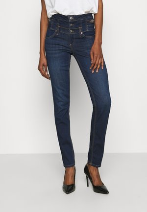 Kvinde iført mørkeblå højttaljede skinny jeans med flere knapper, kombineret med sorte højhælede sko og en hvid top.
