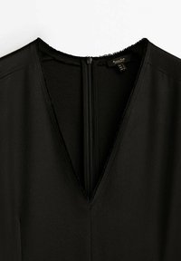 Sort beklædningsgenstand med dyb V-udskæring og glat tekstur. Har en central lynlås og rå kantdetaljer ved udskæringen.