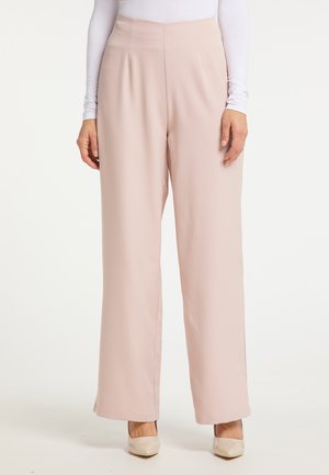 Vrouw die lichtroze, wijde broek met hoge taille draagt, gecombineerd met beige hakken en een witte top met lange mouwen, staand tegen een effen achtergrond.