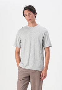 Tricou din bumbac gri, cu mâneci scurte, de tip crew neck, și o croială lejeră. Purtați cu pantaloni bej, pe un fundal alb simplu.
