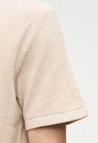 Μπεζ κοντομάνικο πλεκτό τοπ με ορατό υφασμάτινο ράψιμο και διπλωμένη τρέσα, φορεμένο σε χέρι πάνω σε απλό φόντο.