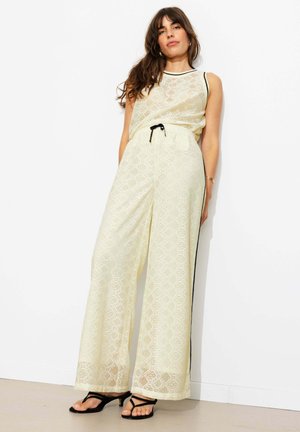 Femme portant un haut sans manches en dentelle crème et un pantalon large avec des bandes latérales noires et des sandales noires, debout contre un mur blanc.
