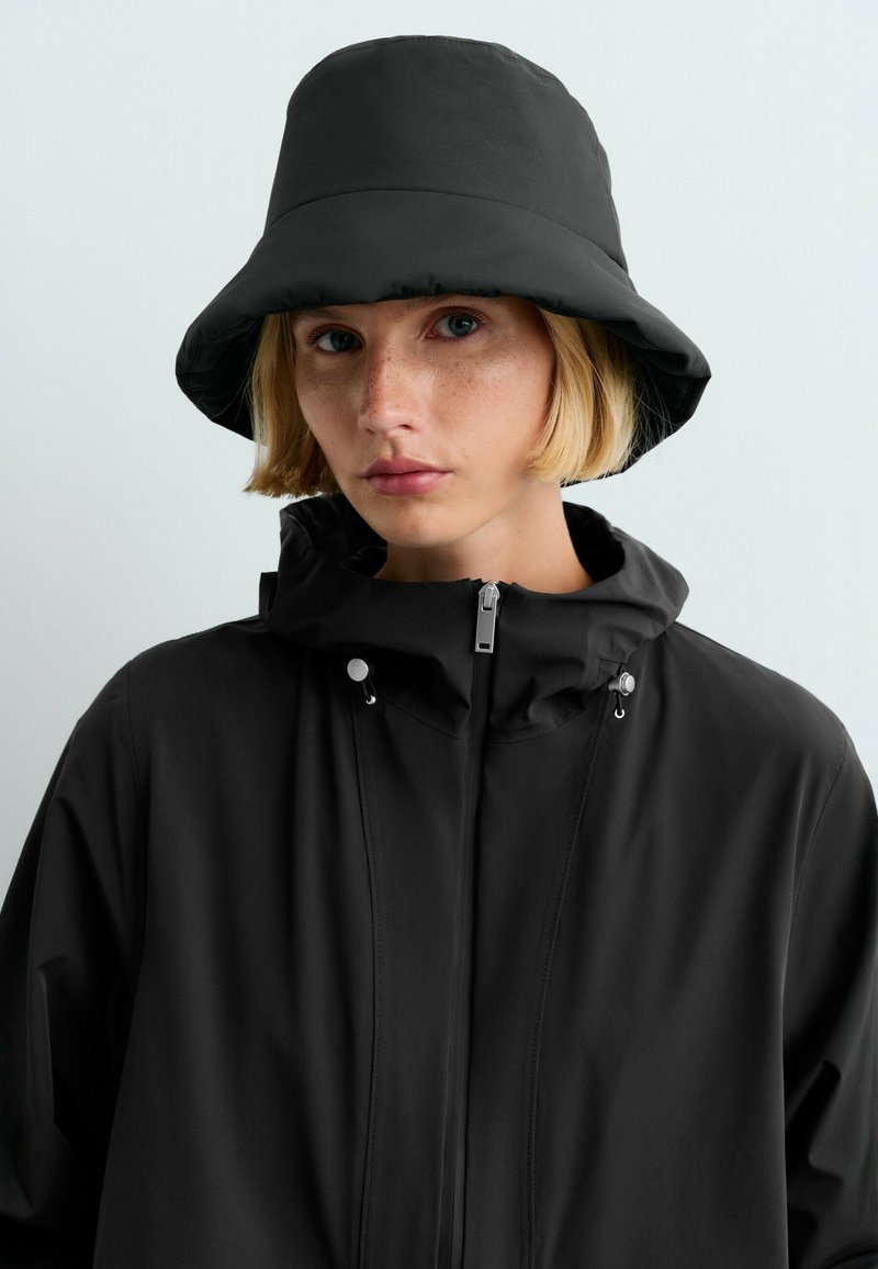Jeune personne aux cheveux blonds courts portant un chapeau bob noir et une veste noire zippée avec cordons sur un fond clair uni.