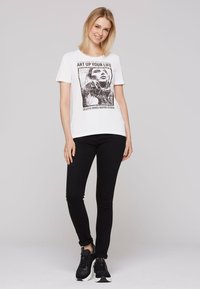 Biała bawełniana koszulka z czarnym nadrukiem graficznym zawierającym tekst i portret, zestawiona z czarnymi dżinsami typu slim-fit i czarnymi sneakersami.