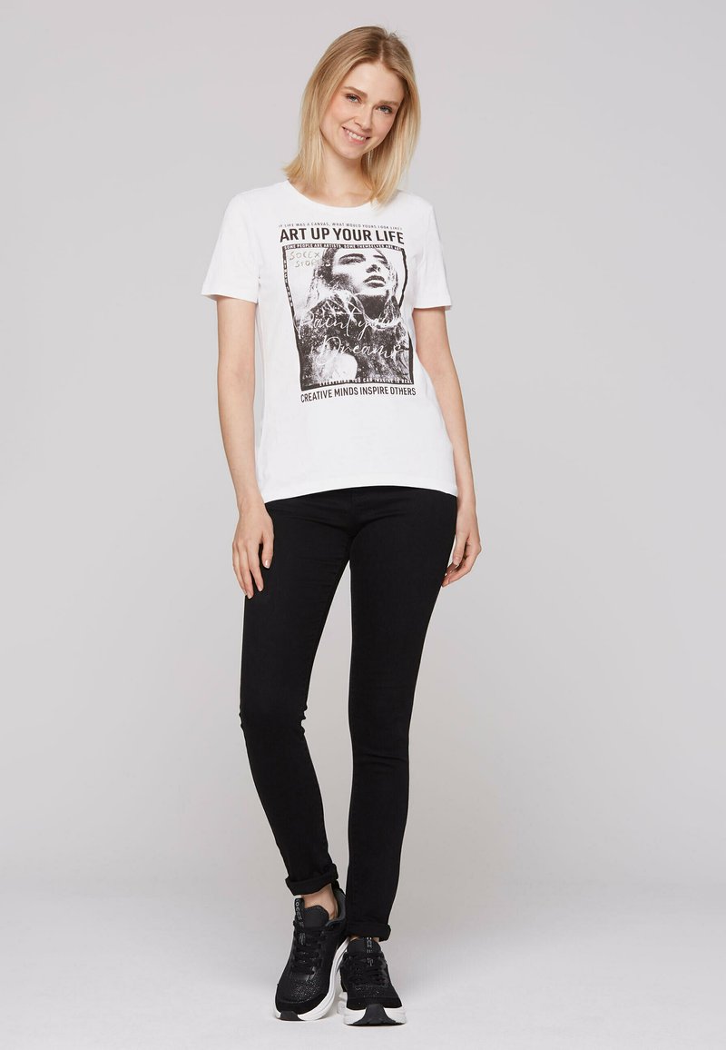 Biała bawełniana koszulka z czarnym nadrukiem graficznym zawierającym tekst i portret, zestawiona z czarnymi dżinsami typu slim-fit i czarnymi sneakersami.