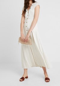 Kobieta w białej sukience midi z guzikami z przodu, w cienkie pionowe paski, trzyma beżową plecioną torebkę i nosi brązowe sandały na odkrytej pięcie.