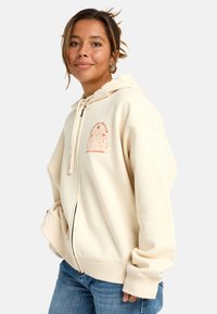 Felpa con zip color crema realizzata in tessuto morbido, con un design grafico sulla parte anteriore, cappuccio con cordino e polsini a coste. Indossata con jeans blu.