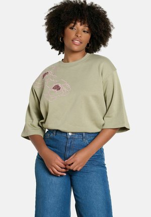 Kobieta o kręconych włosach, ubrana w beżową, oversizową bluzkę z różowym kwiatowym haftem oraz niebieskie, wysokie jeansy, z rękami splecionymi przed sobą.