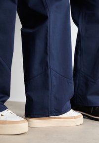 Marineblauwe wijde broek met zichtbare naden, gecombineerd met witte sneakers met een beige zool. Glad stoftextuur.