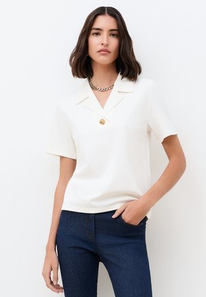 Nainen, jolla on olkapäälle ulottuvat tummat hiukset, valkoinen lyhythihainen paita, jossa on kultainen nappi, ja tummansiniset farkut, käsi taskussa.