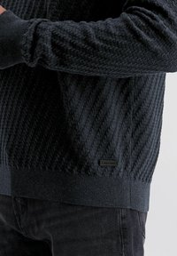 Zwarte gebreide trui met textuur, met ribgebreide manchetten en onderrand, gedragen over een donkere broek, met een klein rechthoekig zwart merketiket nabij de onderrand.