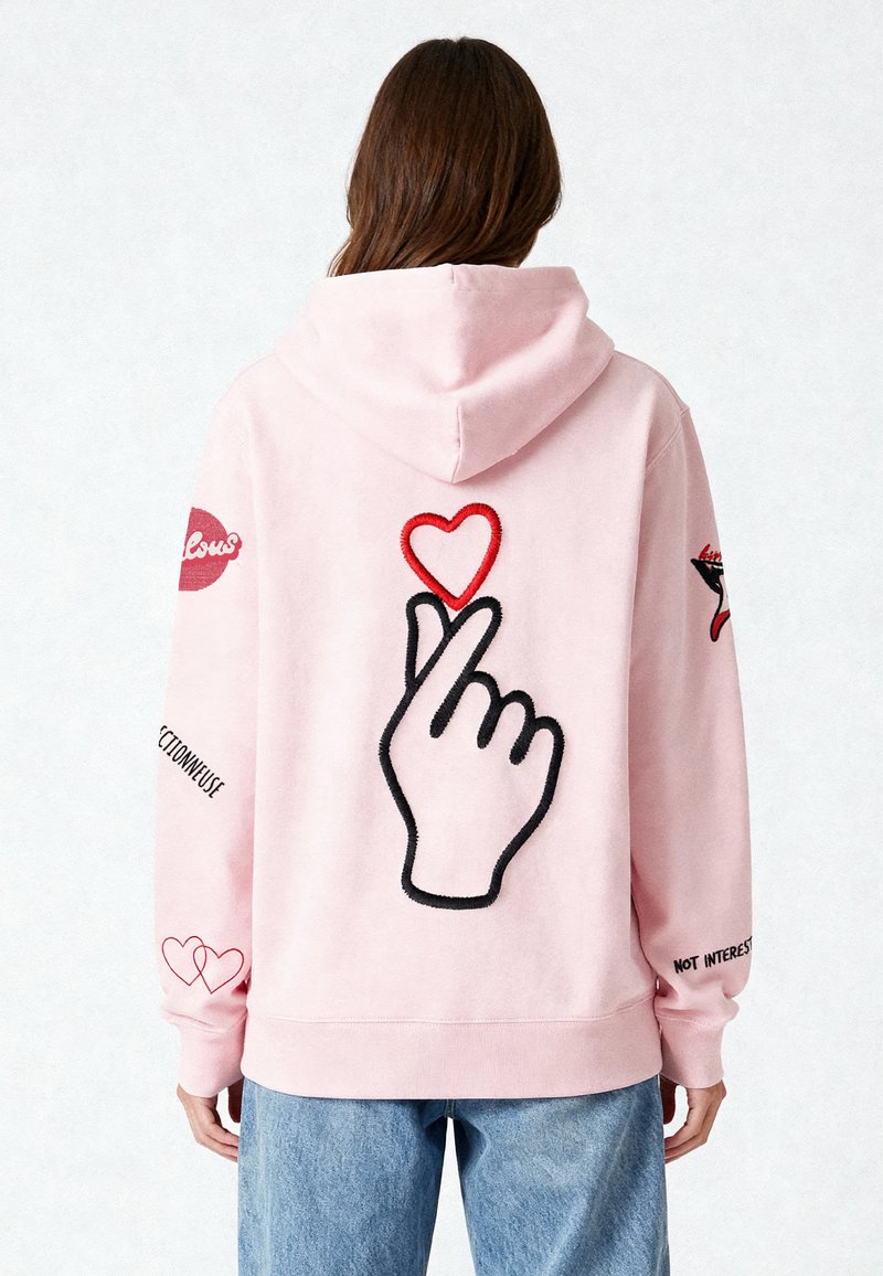Sweat à capuche rose avec un grand design de main noire et un détail de cœur rouge au dos, présentant des broderies sur les manches et une coupe décontractée.