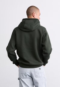 Mørkegrøn hoodie med en stor hætte, ribbede manchetter og kant. Materialet virker blødt og tekstureret. Bæres over lysegrå cargo-bukser.