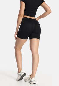 Mujer con shorts biker de talle alto negros y un top corto negro, de pie con una mano en la cadera y zapatillas blancas sobre un fondo blanco.