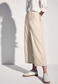 Beige leveled trousers with a high waist, featuring a button closure and side zipper. Paired with white sneakers. Smooth fabric texture.