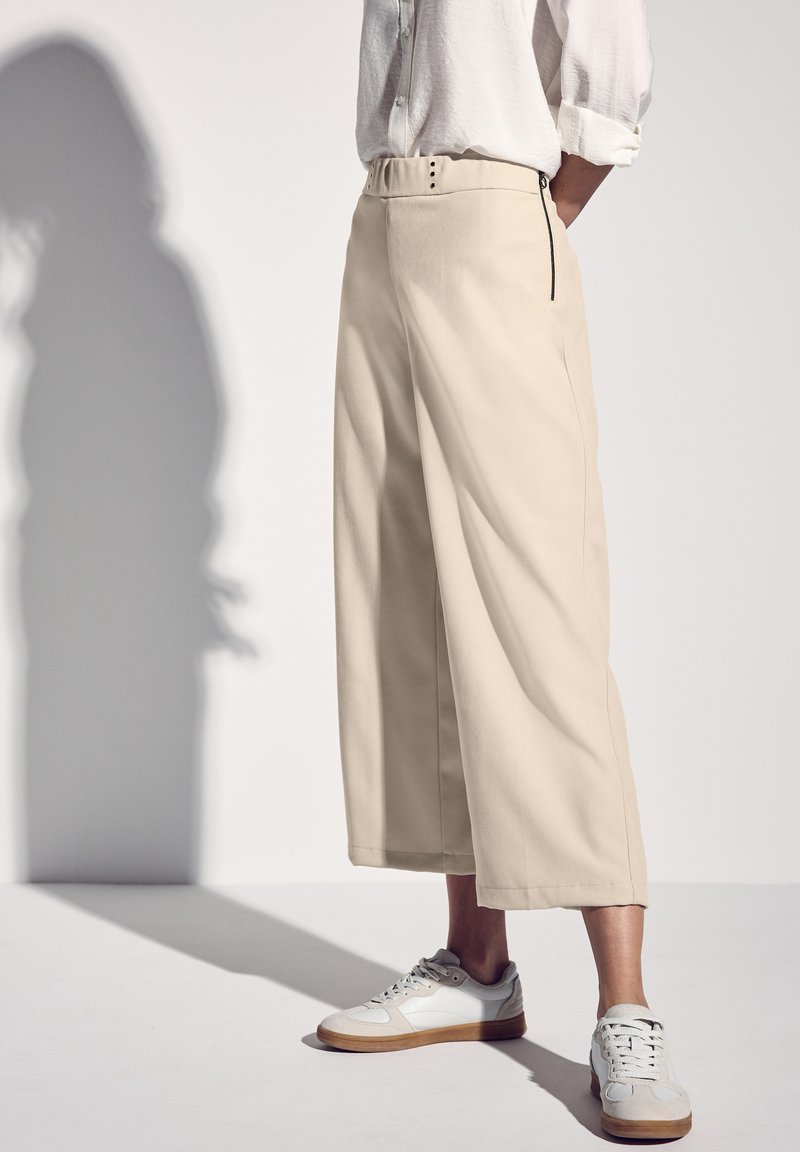 Beige leveled trousers with a high waist, featuring a button closure and side zipper. Paired with white sneakers. Smooth fabric texture.