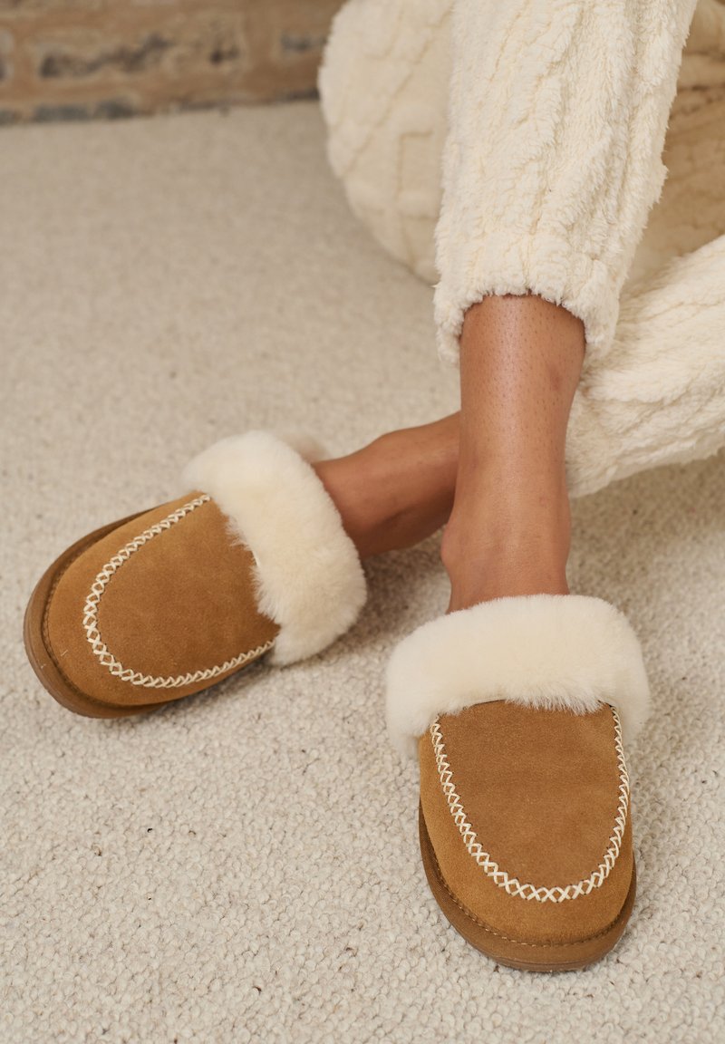 Pantofole in suede marrone con dettagli in pelliccia sintetica, accenti di cucitura bianca, forma arrotondata e suole testurizzate, posate su un tappeto chiaro.