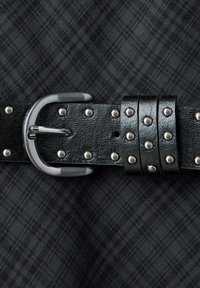 Ceinture en cuir noir avec des clous argentés et une boucle métallique argentée sur un fond en tissu à carreaux gris foncé.
