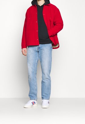 Veste-chemise en polaire rouge portée sur un sweat à capuche noir, associée à un jean bleu clair et des baskets blanches avec des rayures rouges et bleues.