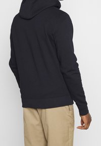 Sudadera con capucha de color azul marino, hecha de tela suave, con mangas largas, un corte relajado y un dobladillo acanalado. Combinada con pantalones de color claro.
