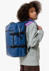 Blå rese-ryggsäck med en slät, glänsande yta. Den har svarta remmar, dragkedjor och metalldetaljer. Användaren har på sig en ljusgrå jacka.