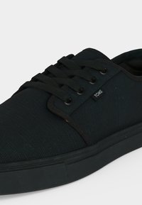 Baskets basses noires avec dessus en tissu, lacets noirs et semelle en caoutchouc, avec une petite étiquette de marque TOMS sur le côté.