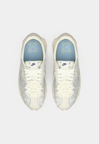 Nike tenisice s gornjim dijelom od svijetlosive mrežaste tkanine, bijelim vezicama, ukrasnim cvjetnim detaljima i plavim ulošcima s geometrijskim uzorkom.