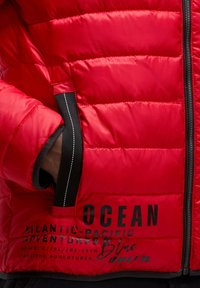 Rote gepolsterte Jacke mit schwarzen Akzenten, sichtbarem Reißverschluss und gedrucktem Branding. Der Stoff wirkt glatt mit einem glänzenden Finish.