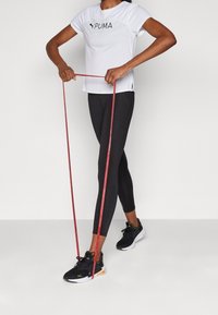 Camisa atlética blanca de manga corta con logo negro, combinada con leggings negros y zapatillas negras. Banda de resistencia roja sostenida en las manos.