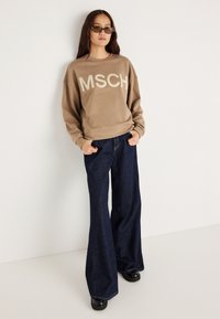 Oversized μπεζ φούτερ με λογότυπο "MSCH", συνδυασμένο με ναυτικό τζιν με φαρδιά γραμμή. Μαύρα chunky παπούτσια και στρογγυλά γυαλιά ηλίου με μοτίβο ολοκληρώνουν το look.