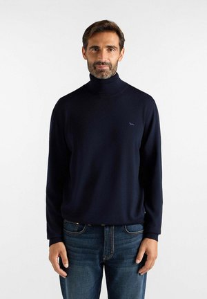 Uomo con capelli corti e scuri e barba indossa un maglione nero a collo alto e jeans blu su uno sfondo bianco uniforme.