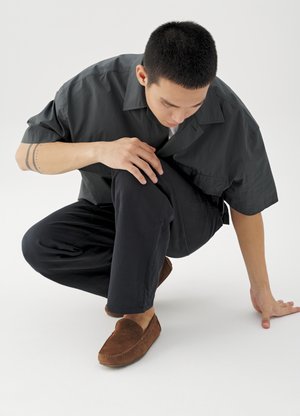 Homme accroupi sur une jambe, portant une chemise sombre, un pantalon noir et des mocassins en daim marron sur fond blanc.