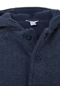 Giacca navy a motivo a spina di pesce con cappuccio foderato in pile, caratterizzata da due bottoni neri e un'etichetta di cura all'interno.