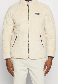 Suddig krämfärgad fleecejacka med hög krage, framzip och sidofickor. Har kontrasterande mörk dragkedja och logotypfäste på bröstet.