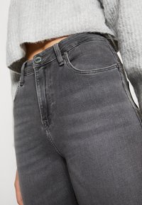 Vaqueros de denim gris con una cintura alta, forma ajustada, textura sutil, cierre de botón metálico y una etiqueta de logo azul en la cinturilla.