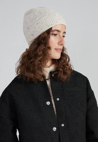 Berretto lavorato a maglia di colore crema con spruzzi, texture a costine. Indossato con una giacca nera a trama con bottoni a pressione argentati.
