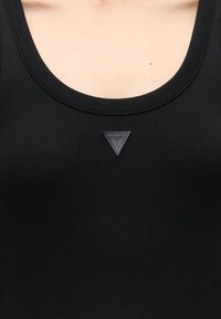 Gros plan d'une personne portant un débardeur noir côtelé avec un petit patch triangulaire du logo Guess sous l'encolure.