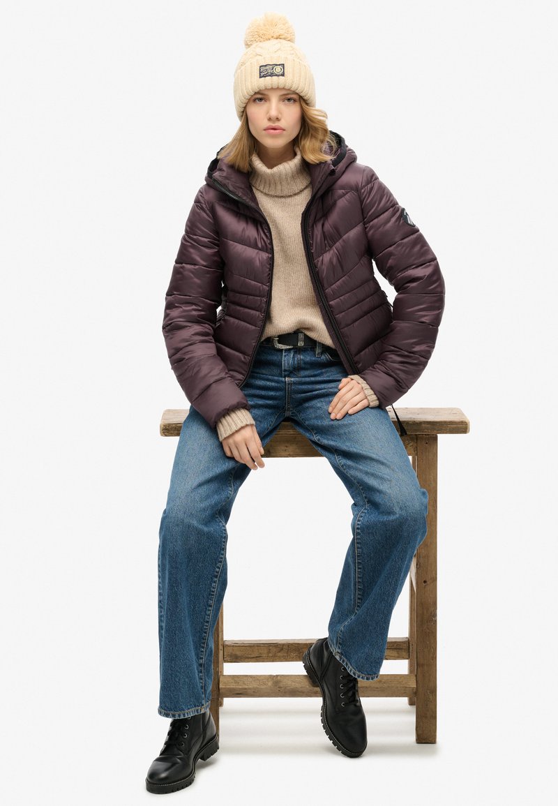 Fioletowa kurtka puchowa z kapturem, beżowy sweter z dzianiny, niebieskie dżinsy i czarne botki za kostkę. Nosi beżową dzianą czapkę z pomponem.