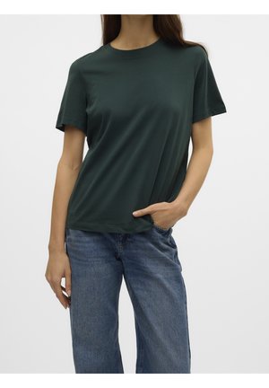 Person iført en mørkegrøn kortærmet t-shirt og blå jeans, stående med den ene hånd i lommen mod en ensfarvet baggrund.