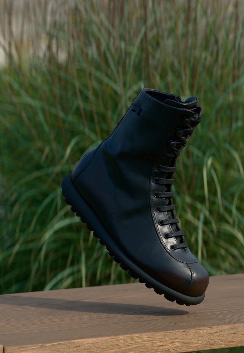 Botte en cuir noir à lacets suspendue au-dessus d'une surface en bois avec de hautes herbes vertes en arrière-plan flou.