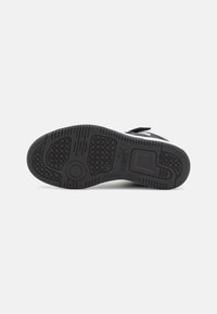 Schwarze Sportschuhsohle mit einer strukturierten Gummioberfläche, die kreisförmige und sechseckige Muster aufweist, sowie einem glatten Fersenbereich und einem Markenlogo.