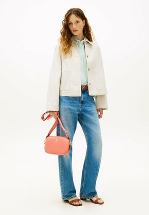 Jeune femme aux longs cheveux bouclés portant une veste crème, une chemise bleu clair, un jean bleu, des sandales marron et tenant un sac bandoulière corail.