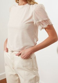 Top corto a maniche corte beige chiaro con maniche a sbuffo trasparenti e bordo in pizzo, abbinato a jeans bianchi in denim a vita alta con tasche laterali.