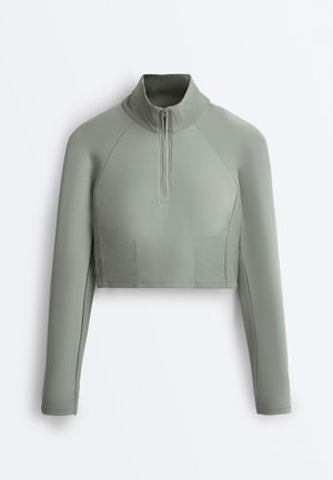 Langärmelig cropped sporttop in saliegroen met geribbelde col en zijpaneel details, voorzien van een halve ritssluiting aan de voorkant.