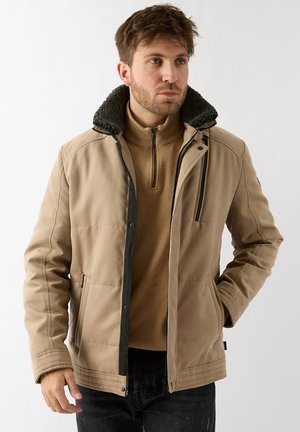 Man draagt een beige jas met een zwarte fleecekraag en een beige trui met een kwart rits, kijkt naar rechts met één hand in de zak tegen een witte achtergrond.