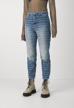 Person trägt hochtaillierte blaue Jeans mit Muster, beige Schnürstiefel und ein langärmeliges Netzshirt mit einem schwarzen Crop-Top darunter.