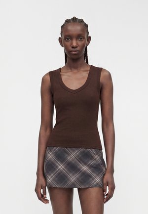 Woman with braided hair wearing a sleeveless brown top and a black and white plaid mini skirt, standing against a plain white background.